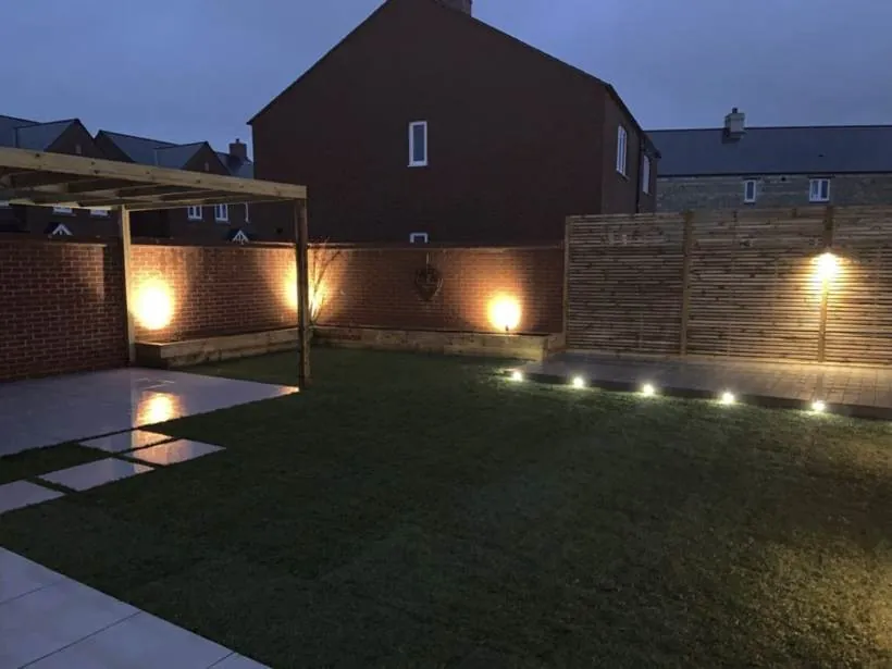 Garden at dusk with warm uplights on a brick wall, LED deck lights and a fence spotlight, Chesterton near Bicester