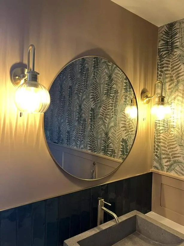 Two ribbed glass wall lights either side of a round brass mirror in a bathroom with fern wallpaper, Bicester