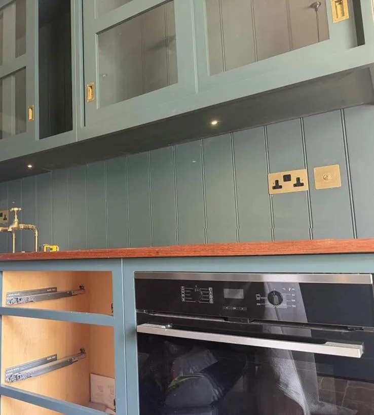 Antique brass double sockets and switch on green panelled kitchen wall above a timber worktop, Kiddington