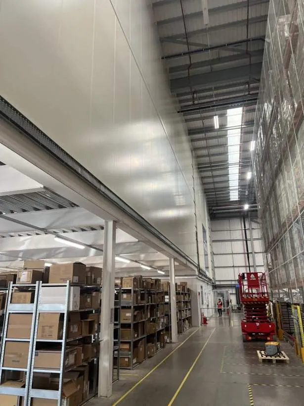 Tall warehouse aisle with new high-level cable tray along the mezzanine edge and a scissor lift in use, Bicester