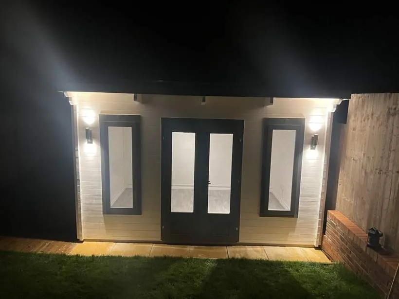 Garden log cabin at night lit by outdoor wall lights either side of the doors, with internal lighting and power, Faringdon, Oxfordshire