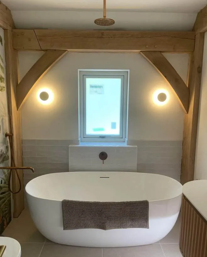 Finished bathroom with two circular wall lights either side of a window framed by oak beams, above a freestanding bath, village near Bicester