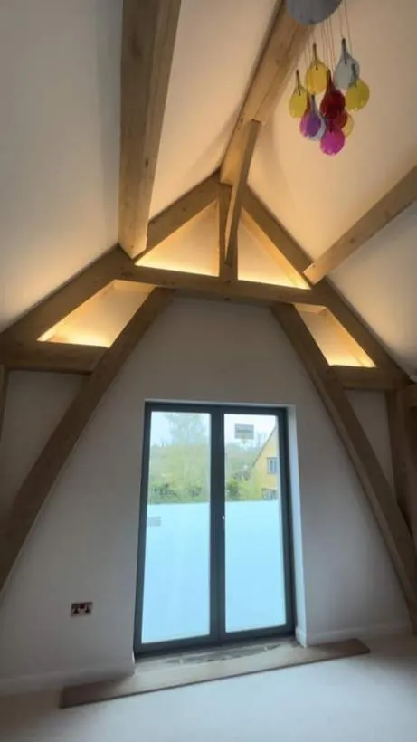 Vaulted oak frame room with warm LED strip lighting hidden along the apex timbers and a colourful glass cluster pendant, village near Bicester