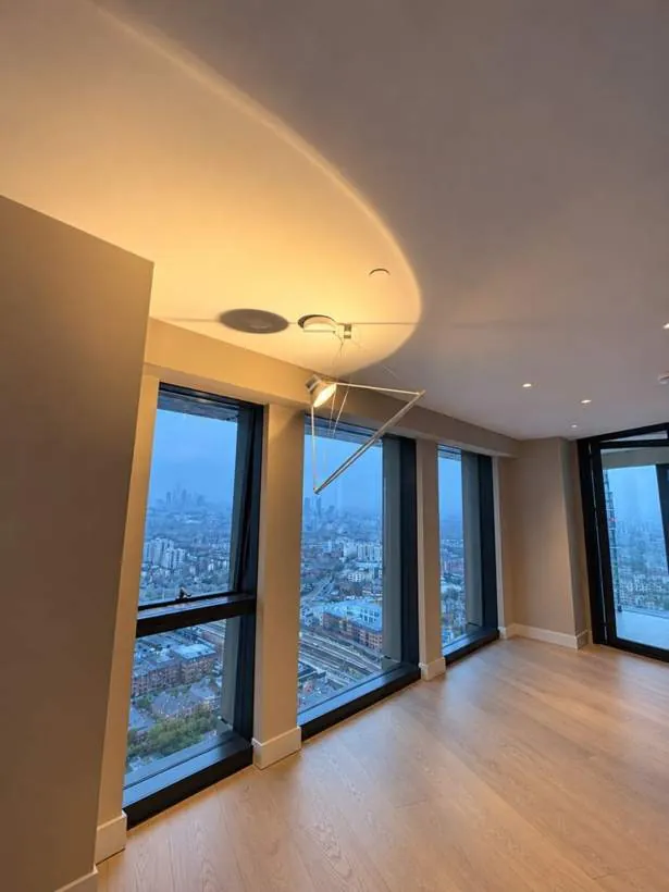 Architectural suspended spotlight pendant washing a curved ceiling with warm light, beside LED downlights and floor-to-ceiling windows overlooking London, Vauxhall
