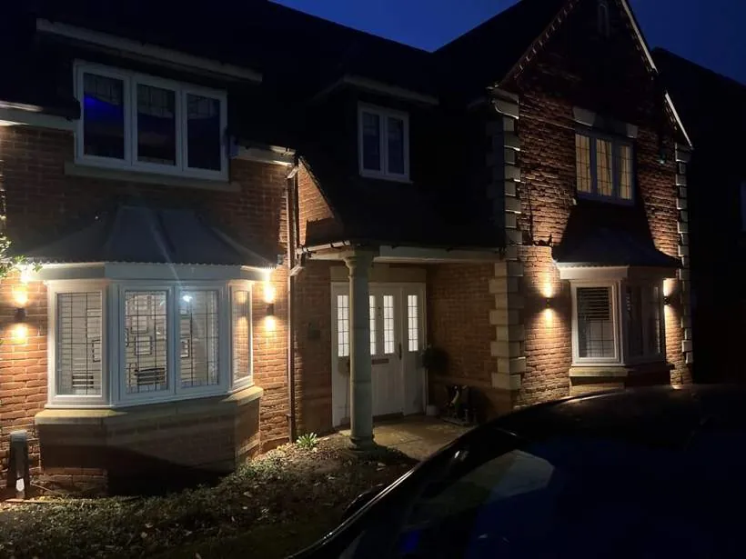 Four up and down wall lights washing light across the brick front of a detached house at dusk, Bicester